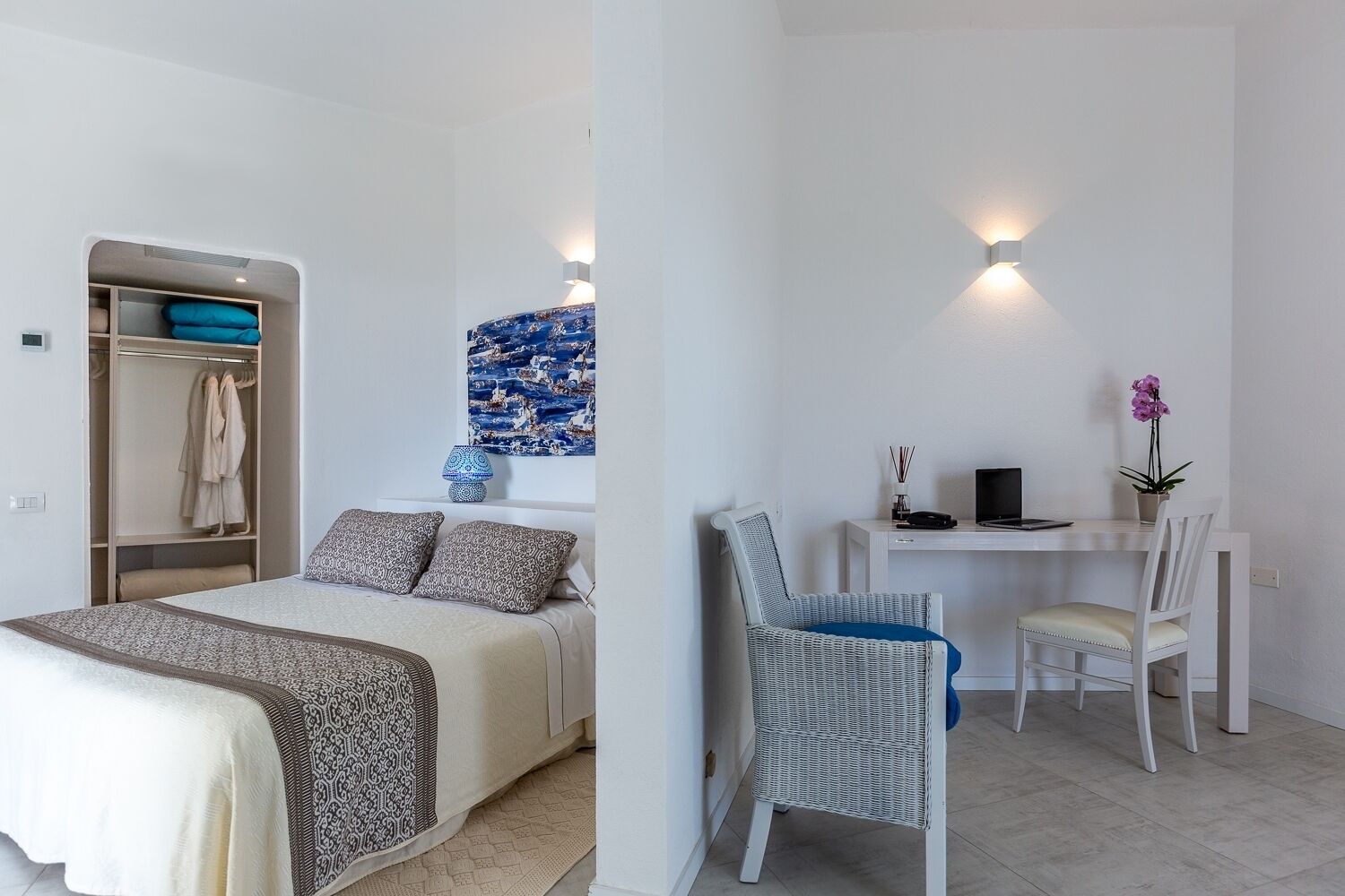A bedroom with a bed, a wardrobe, a desk with a chair, and a wall-mounted lamp.