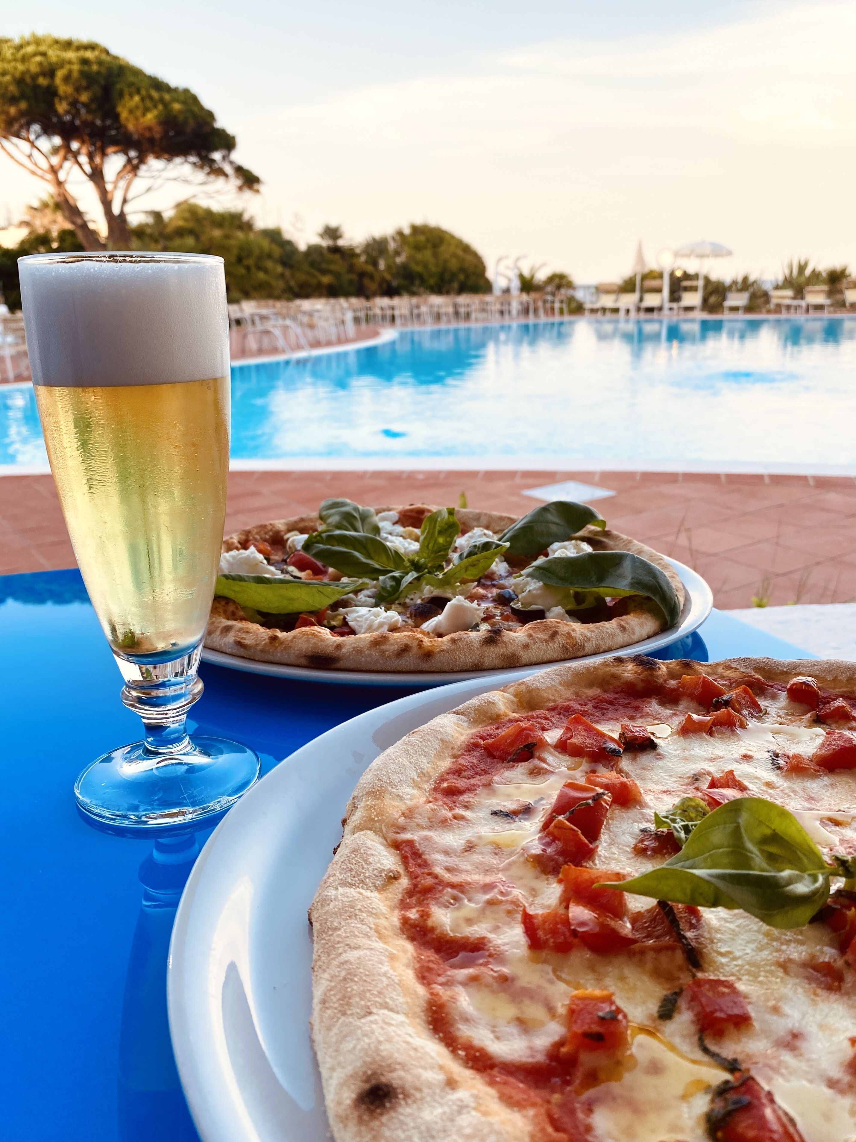 Ein Esstisch mit einem Bierglas, zwei Pizzen und einem Pool im Hintergrund.