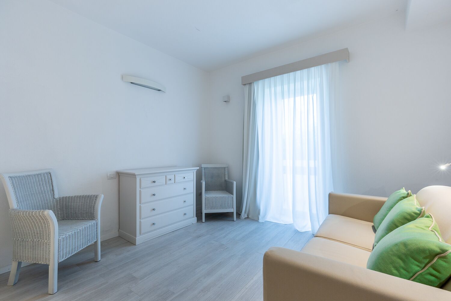 Una habitación luminosa y minimalista con un tocador blanco, una silla de mimbre, una cuna pequeña y un sofá beige con cojines verdes.