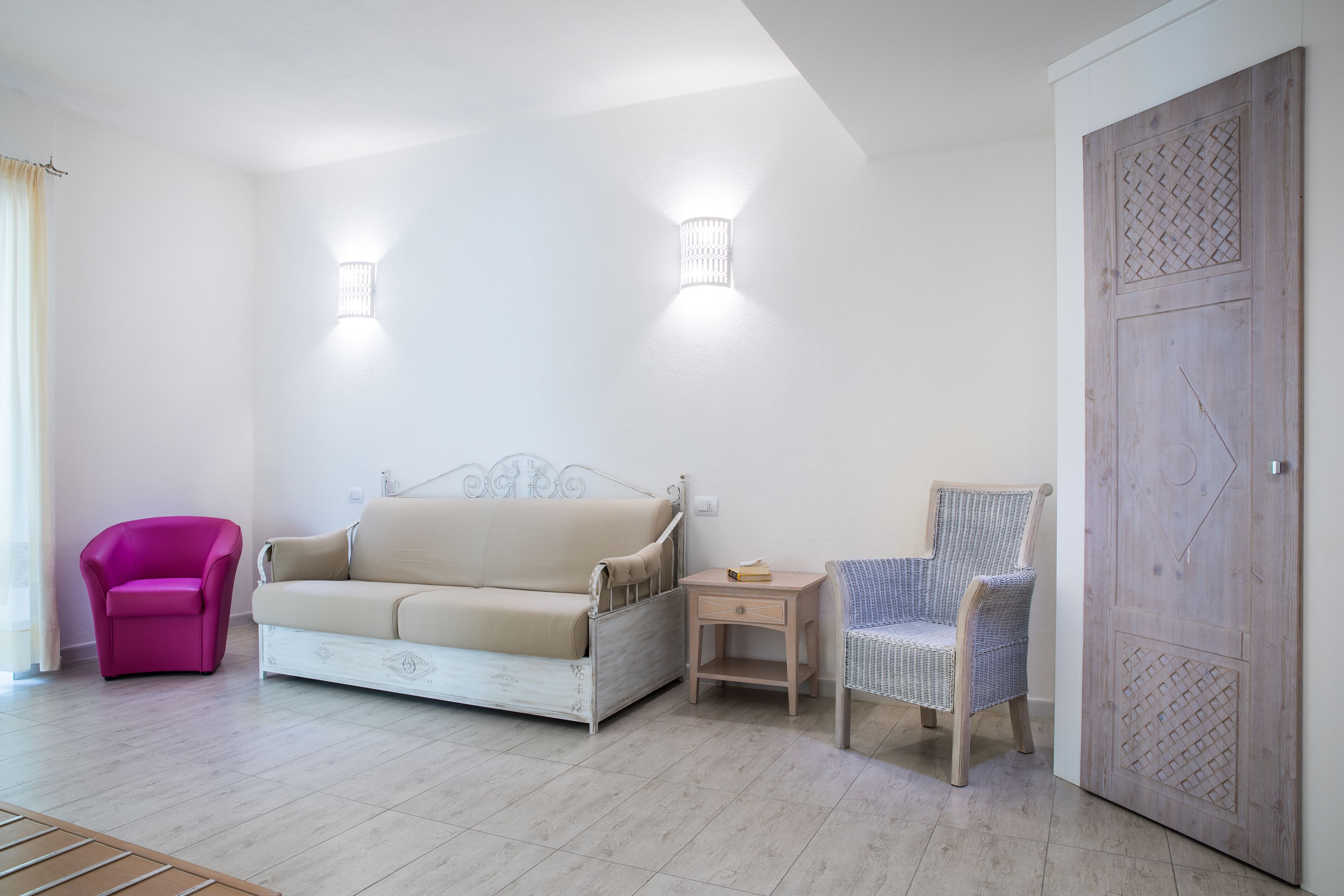 A bedroom with a bed, a pink chair, a beige sofa, a wooden nightstand, and a wardrobe.