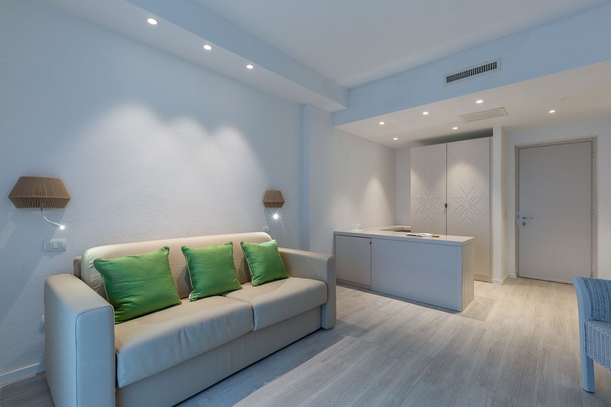 A modern living room with a sofa, a small kitchen area, and a door leading to another room.