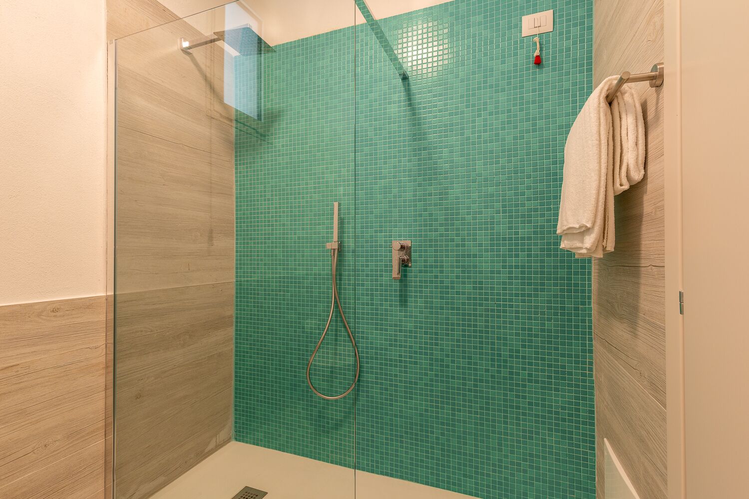 A glass-enclosed shower with a green tiled wall, a towel rack with a white towel, and a wooden panel on the left.