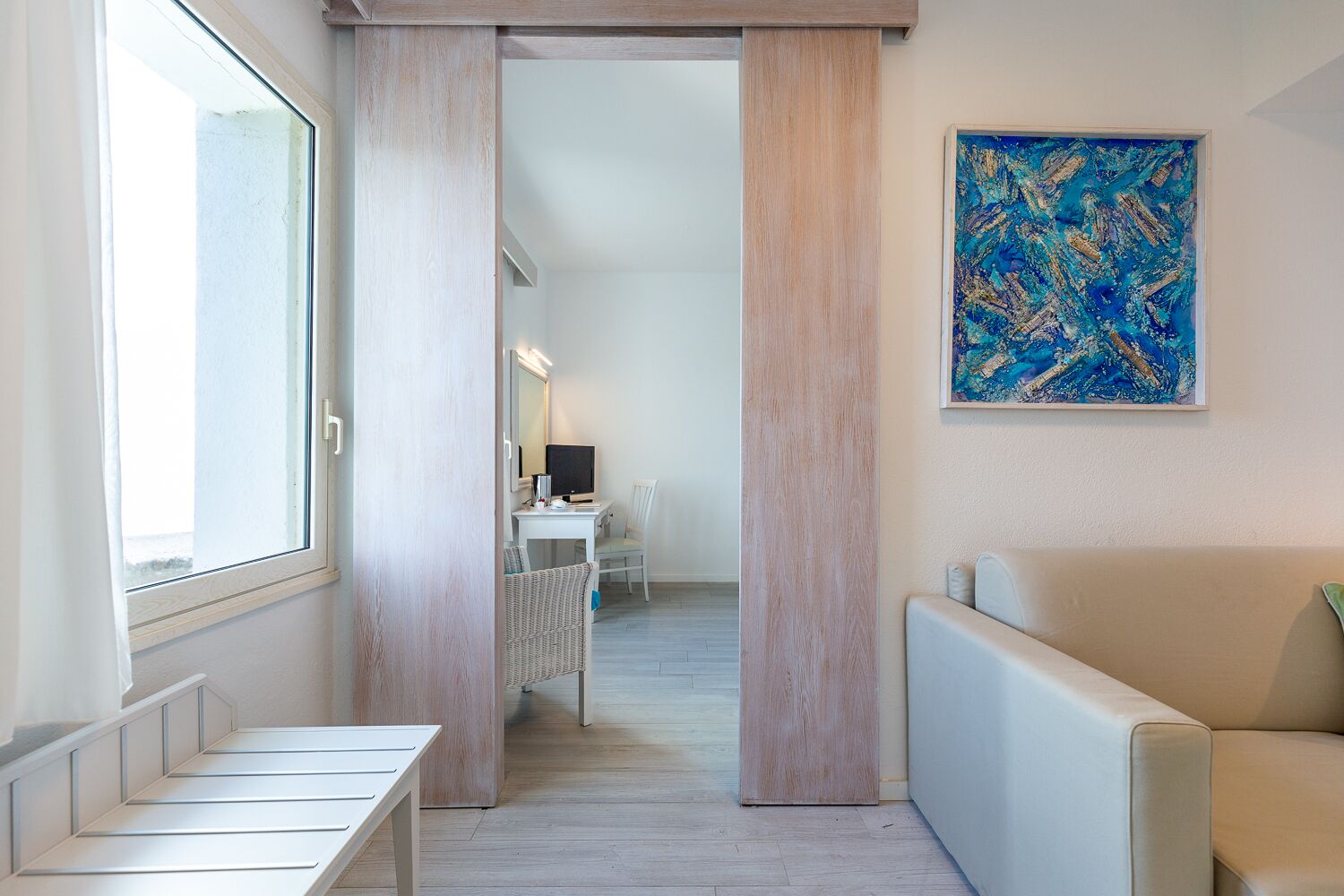 A modern hotel corridor with a seating area, a desk with a computer, and a large abstract painting on the wall.