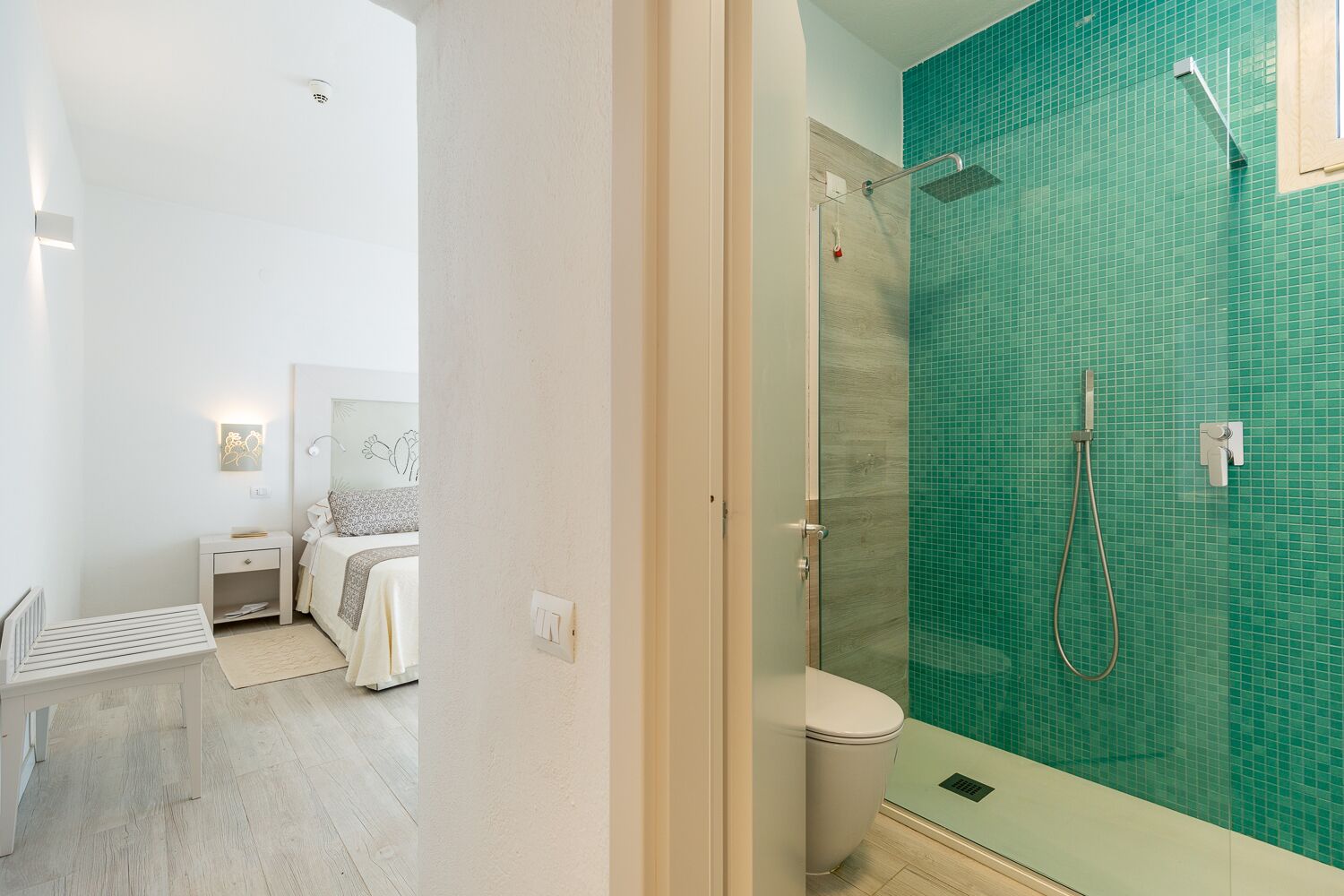 Ein modernes Badezimmer mit Duschkabine aus Glas, Toilette und Nachttisch, im Hintergrund ein Bett.