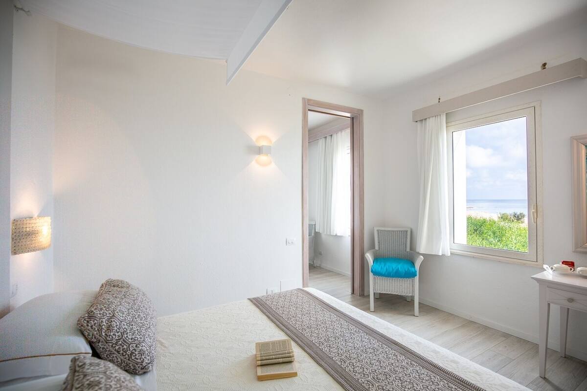 A bedroom with a bed, a chair, a desk, a mirror, and a window with a view of the sea.
