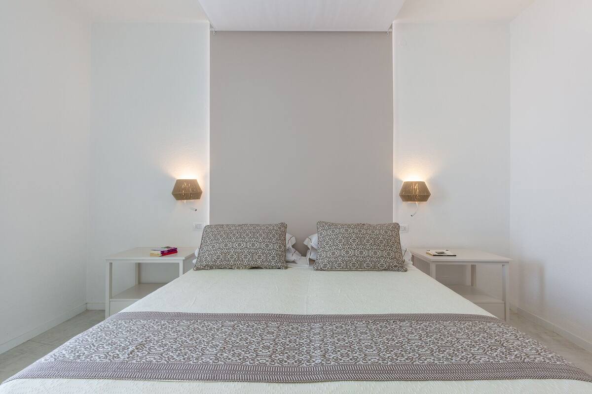 A minimalist bedroom with a large bed, two bedside tables, and wall-mounted lamps.