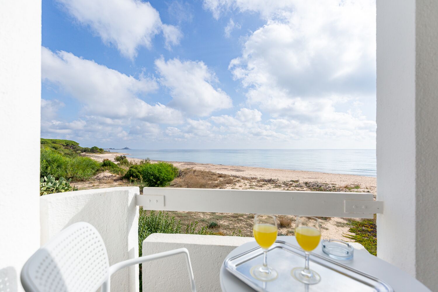 A balcony with a table, two glasses of yellow liquid, and a view of a beach and ocean.