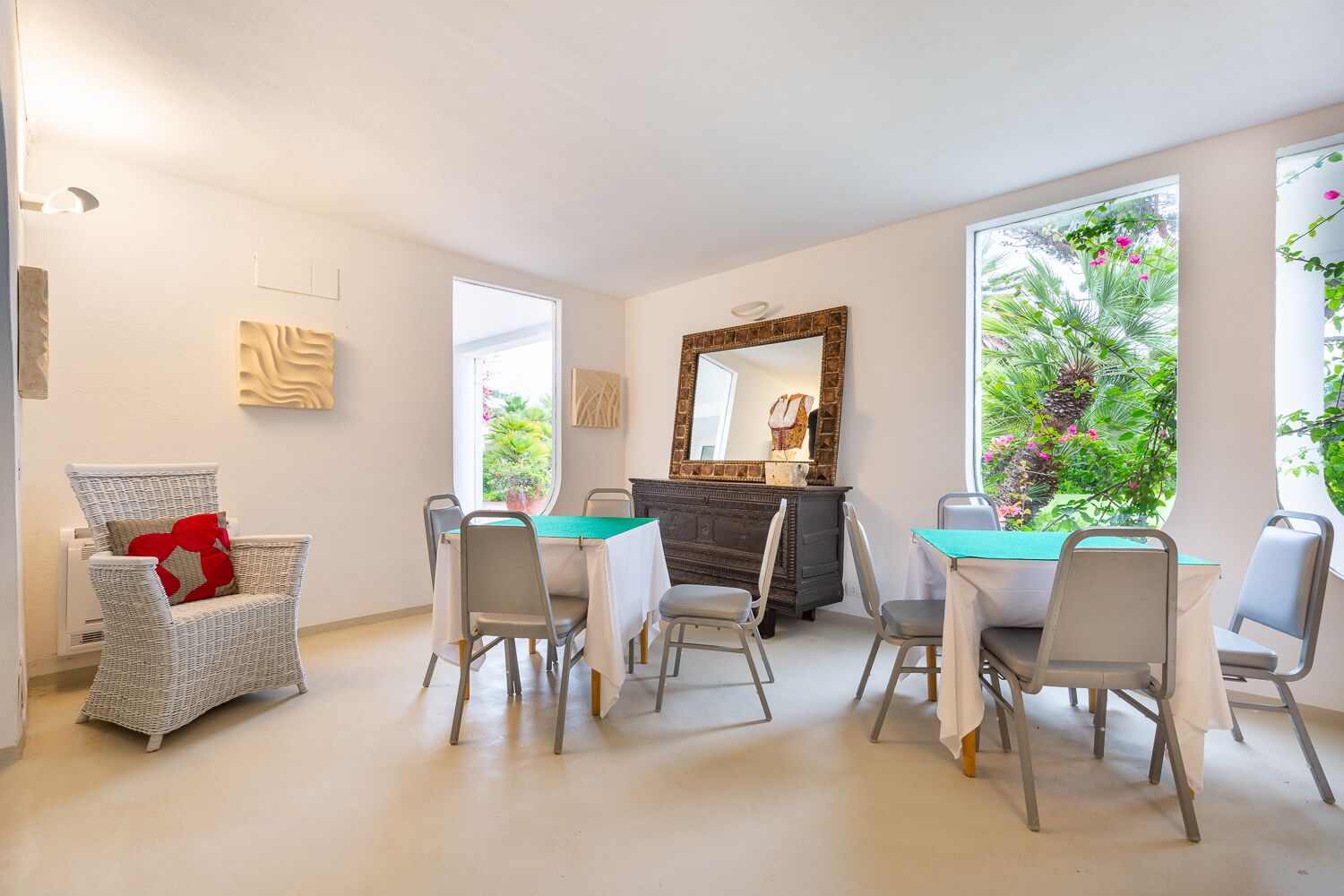 Un comedor con una mesa y sillas, una silla de mimbre con un cojín rojo, un armario de madera oscura y un espejo grande en la pared.