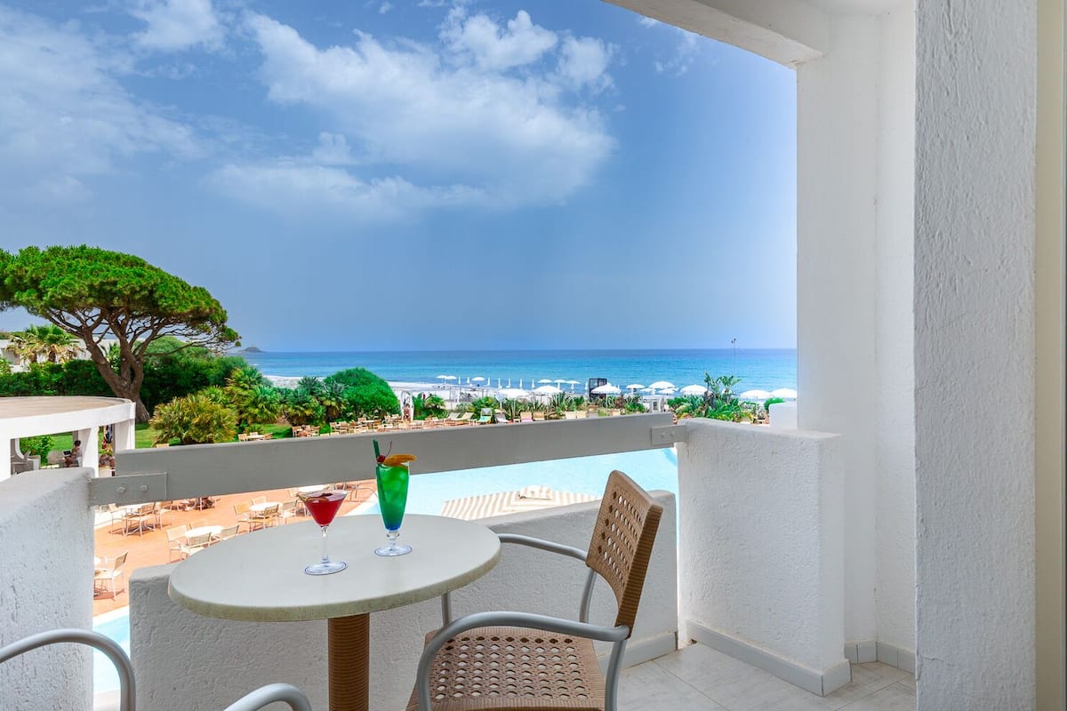 A balcony with a table and chairs, overlooking a beach and pool area.