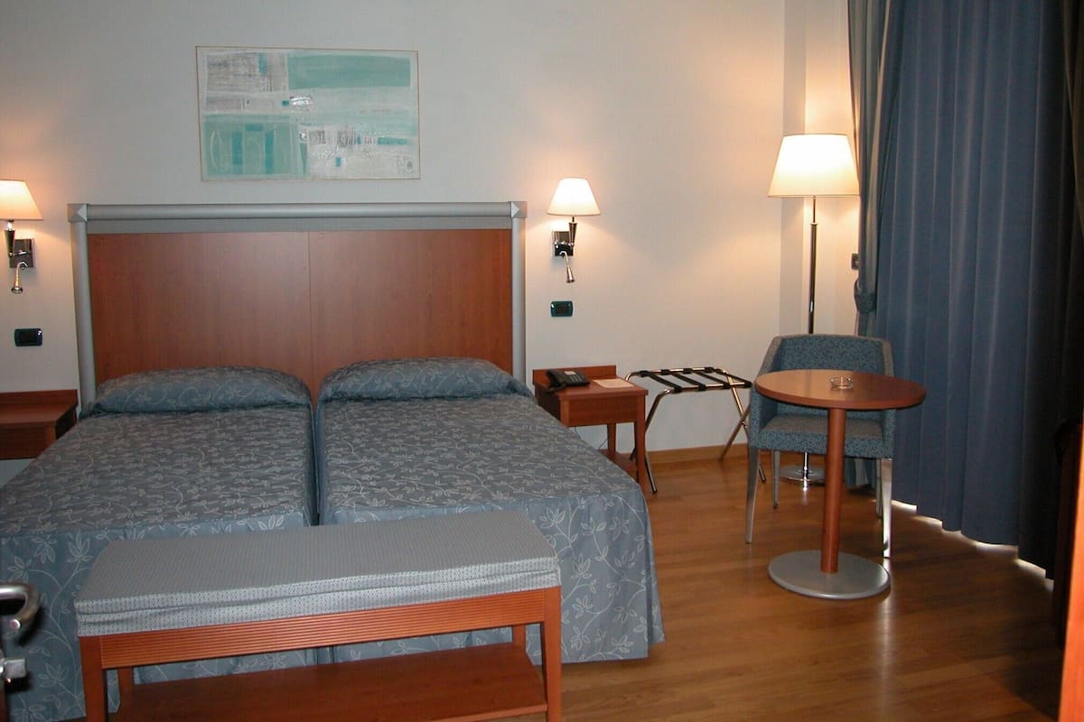 A hotel room with a double bed, a small desk, a chair, a bedside table, and a lamp.