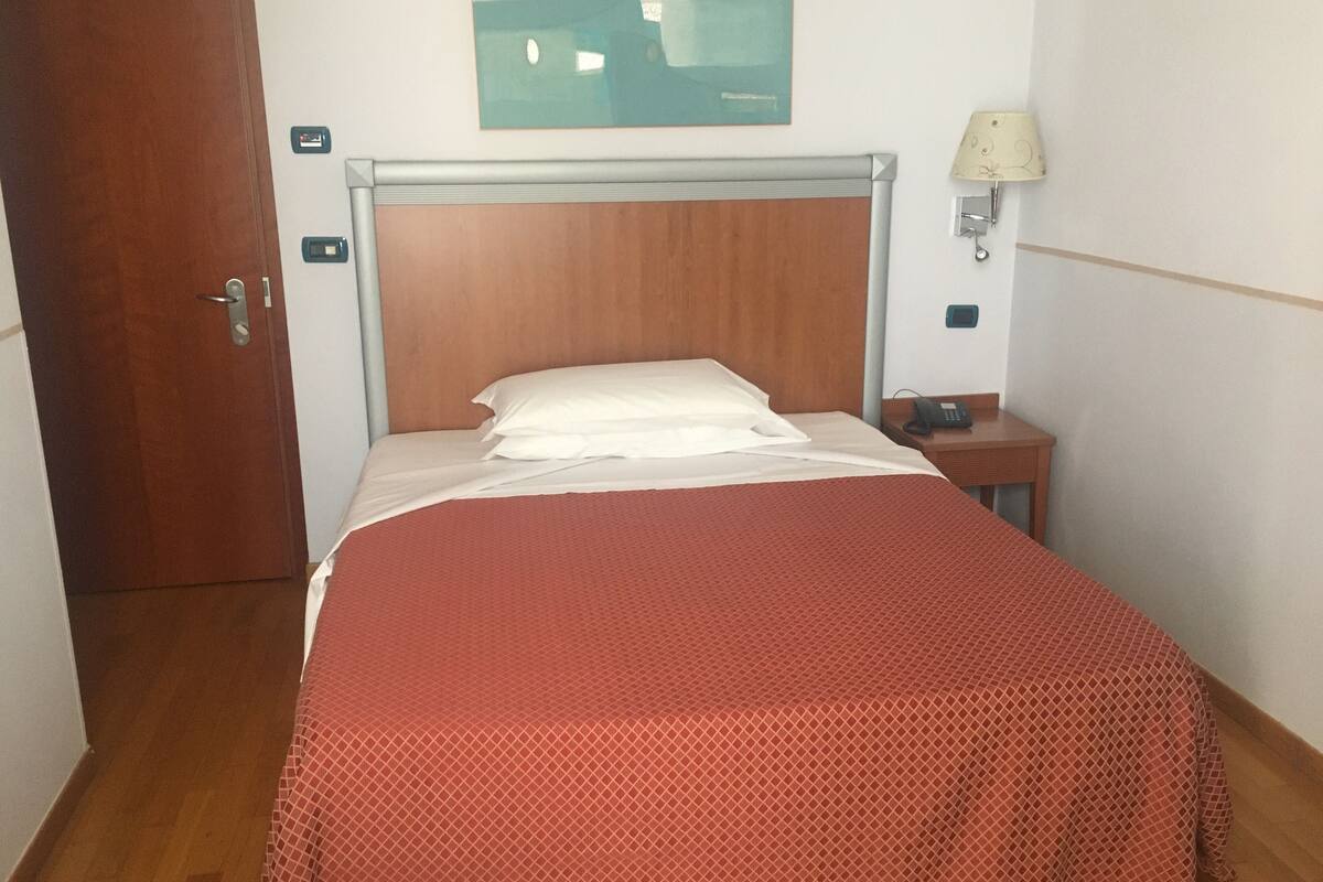 A hotel room with a wooden headboard, a bed with a red patterned bedspread, a bedside table with a lamp, and a wall-mounted phone.