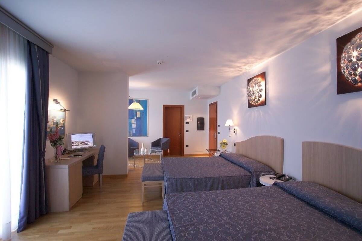 A hotel room with two beds, a desk with a computer, a chair, and a small table with a lamp.