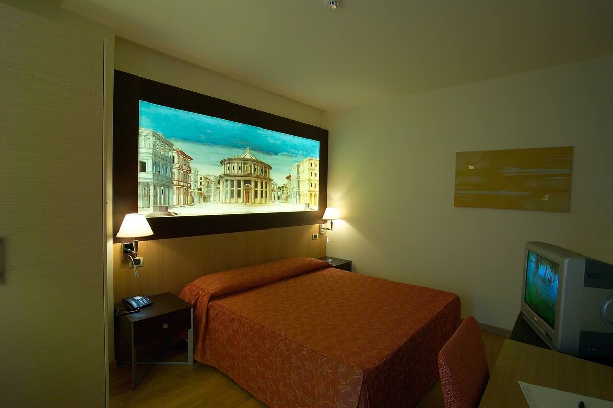 A hotel room with a bed, a desk with a computer, a lamp, and a painting of a cityscape.