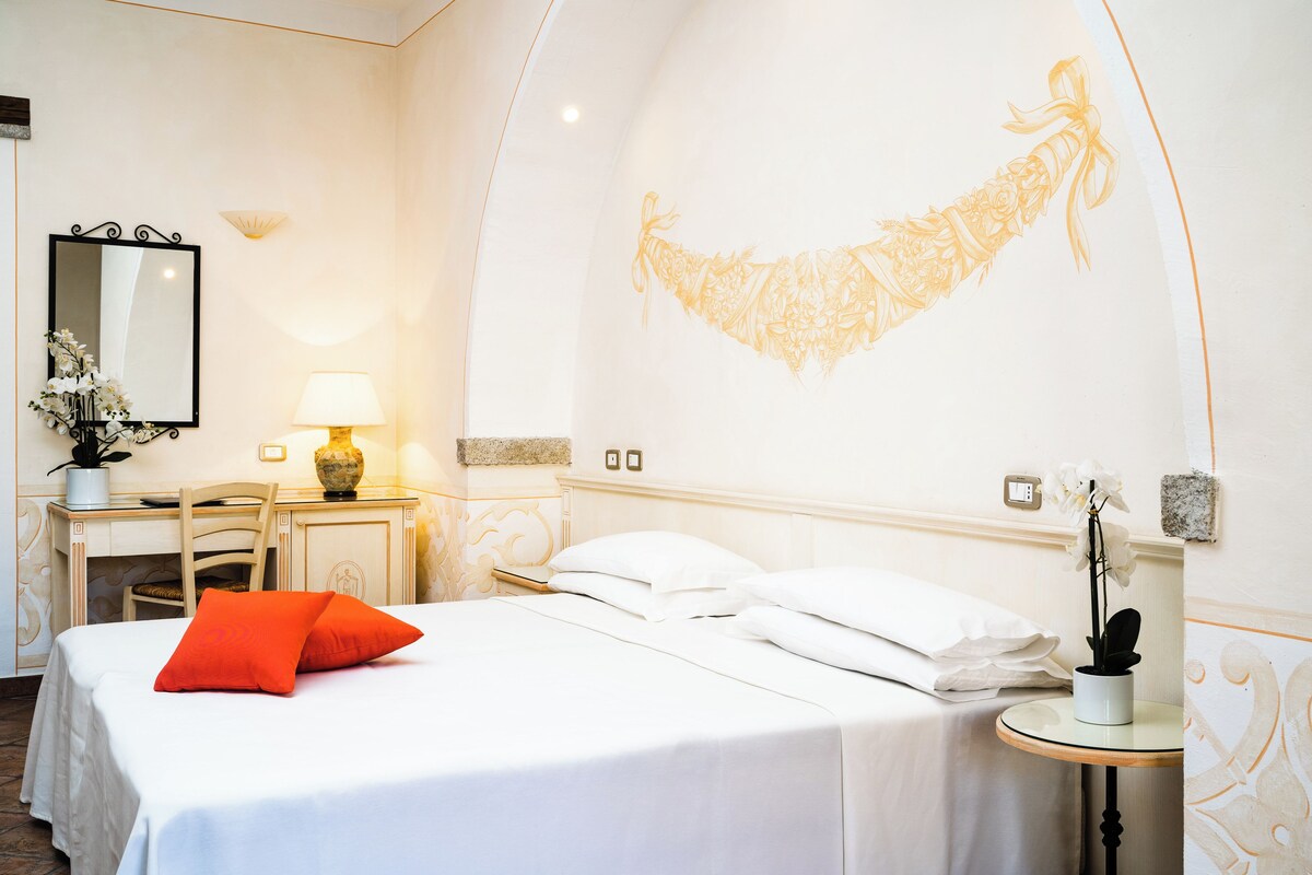 A hotel room with a bed, a desk, and a chair. There is a lamp on the desk and a vase with flowers.