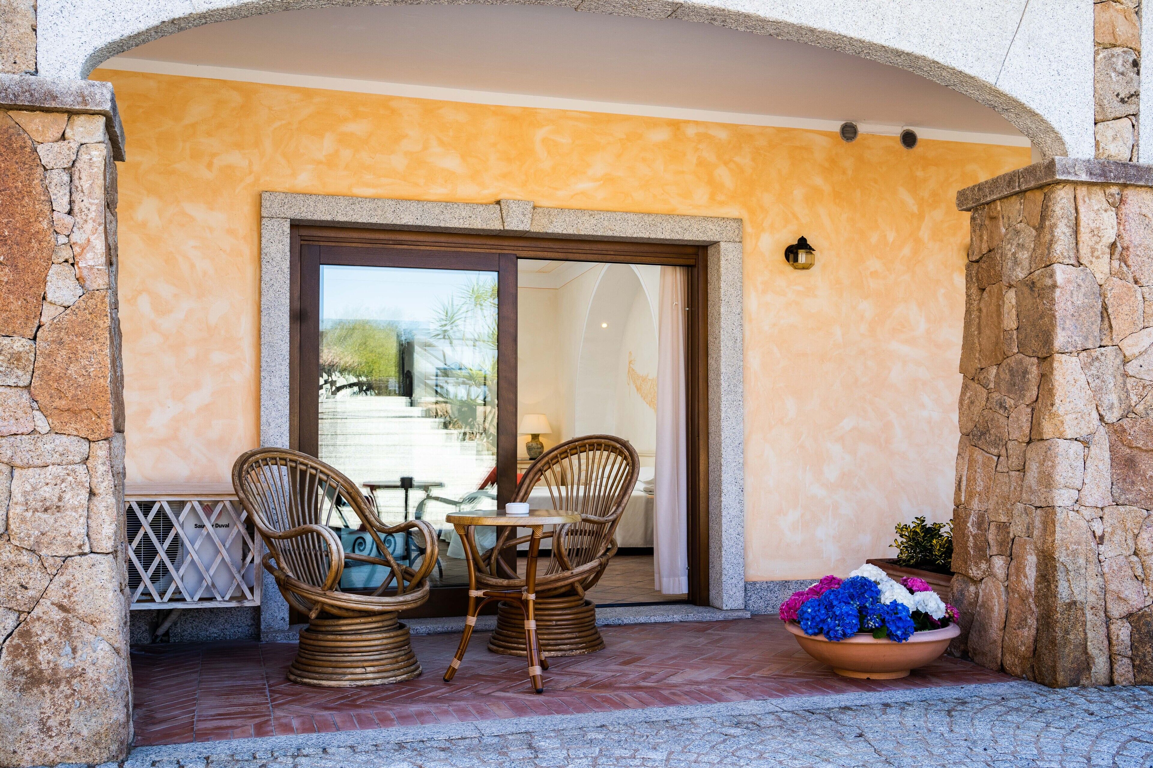 An outdoor seating area with wicker chairs and a table, a potted plant with flowers, and a stone wall.