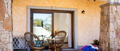 An outdoor seating area with wicker chairs and a table, a potted plant with flowers, and a stone wall.