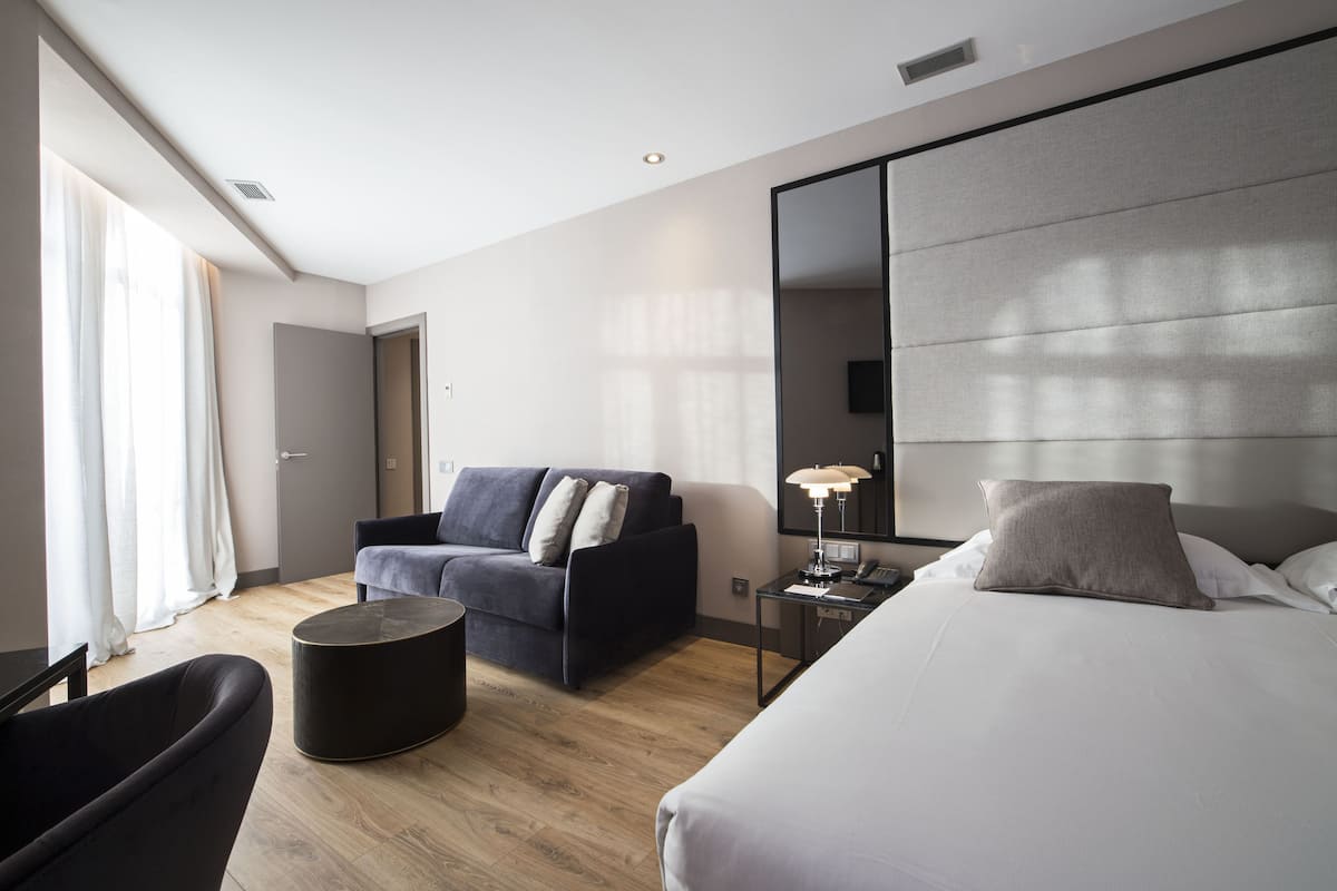 A modern hotel room with a bed, a sofa, a round ottoman, a nightstand with a lamp, and a large mirror.