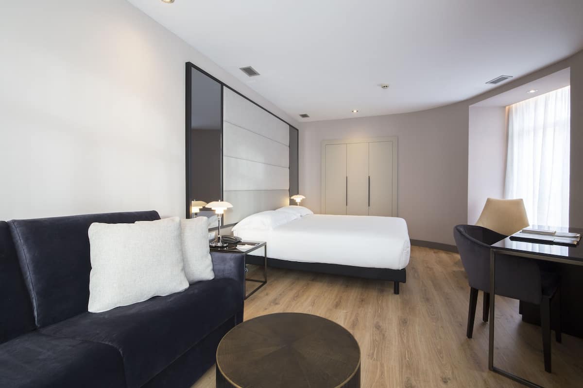 A modern hotel room with a bed, a sofa, a desk, and a large mirror.