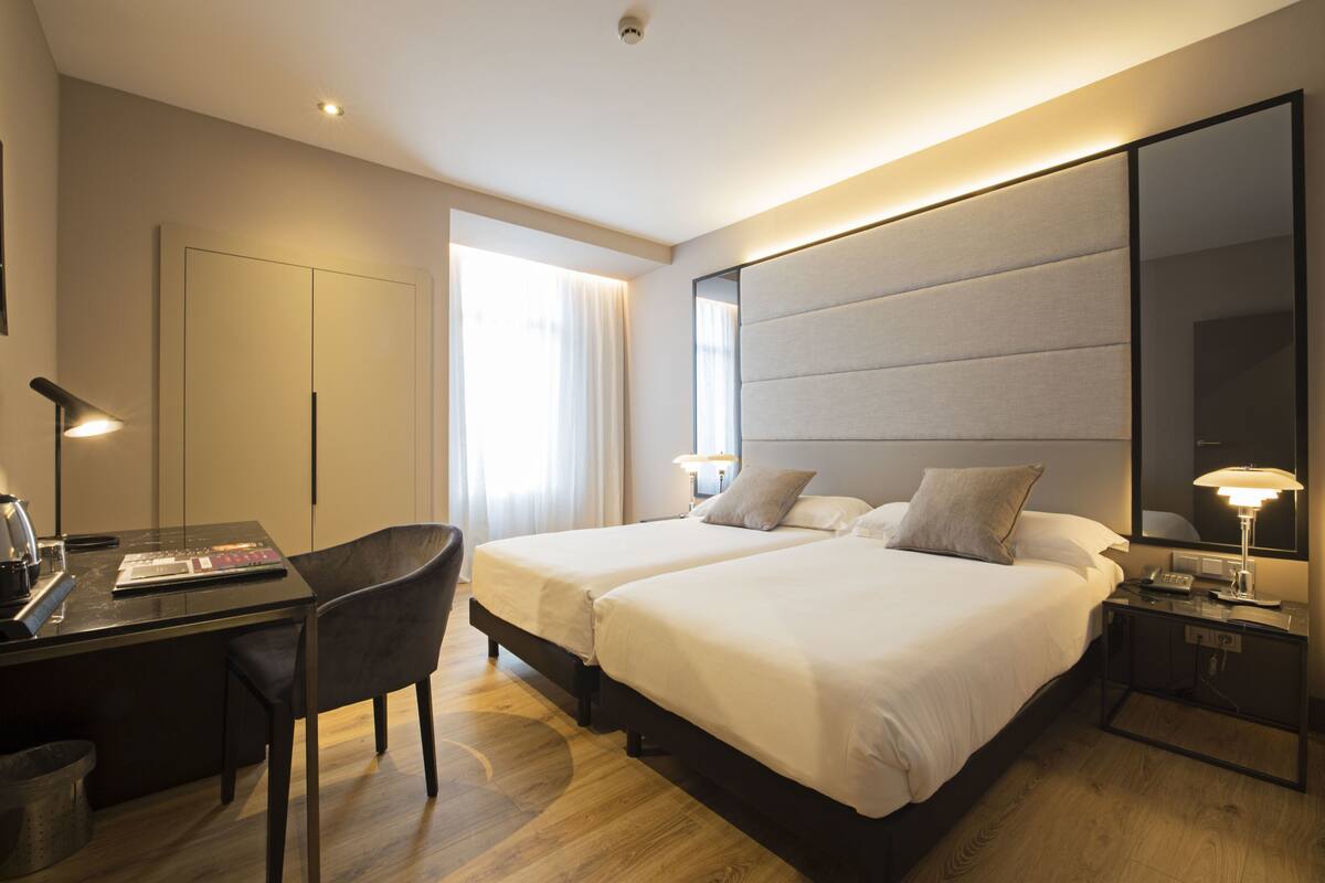 A hotel room with two beds, a desk, a chair, a large mirror, and a nightstand with a lamp.
