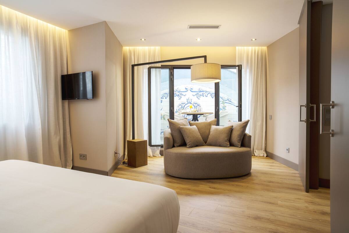 A modern hotel room with a large bed, a round seating area, a flat-screen TV, and a view of a balcony with a railing.