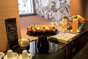 A buffet setup with a variety of food items, a sign for 'Zenit Hotels', and a window with a view of buildings in the background.