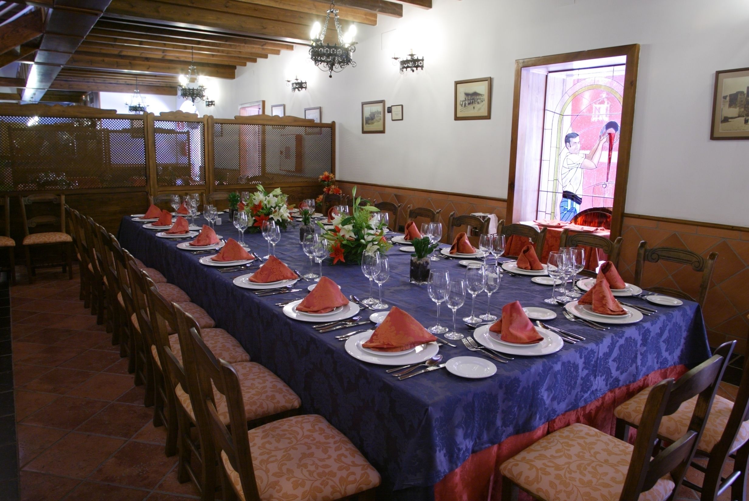 Une salle à manger avec une longue table dressée pour le repas, des chaises en bois et une fenêtre décorative avec un vitrail.