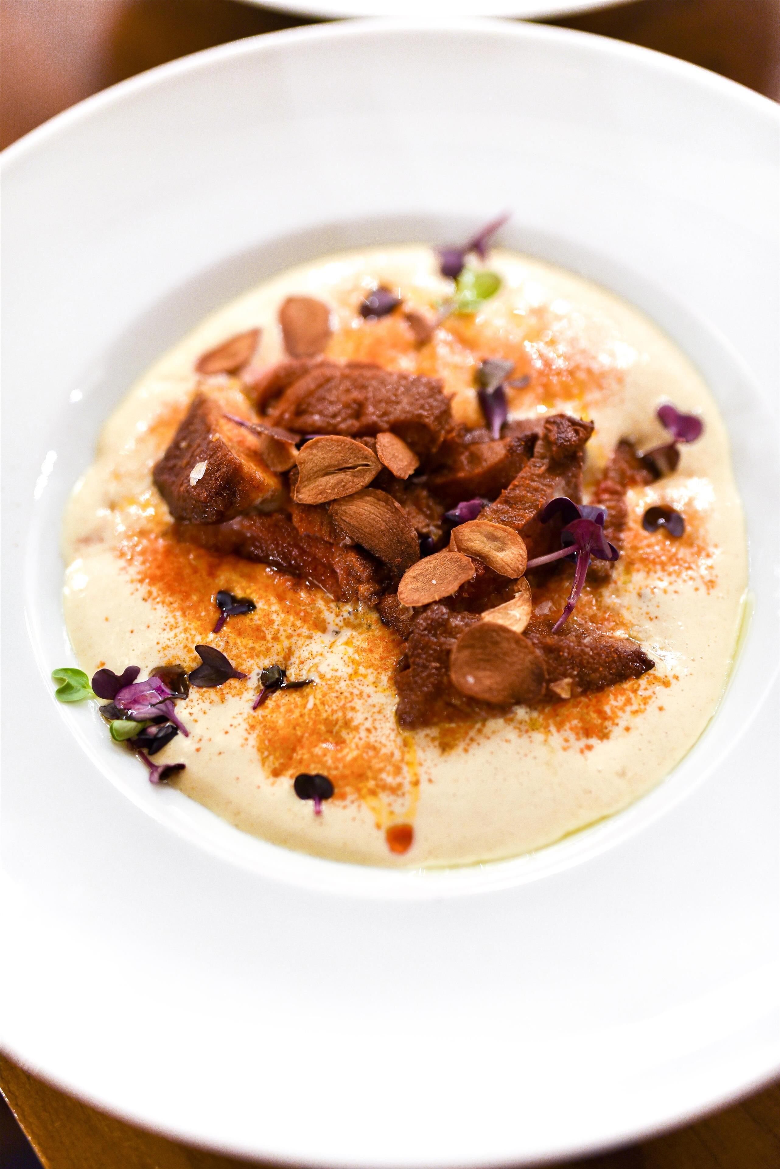 Un plato blanco con un postre cremoso cubierto de almendras y flores comestibles moradas.