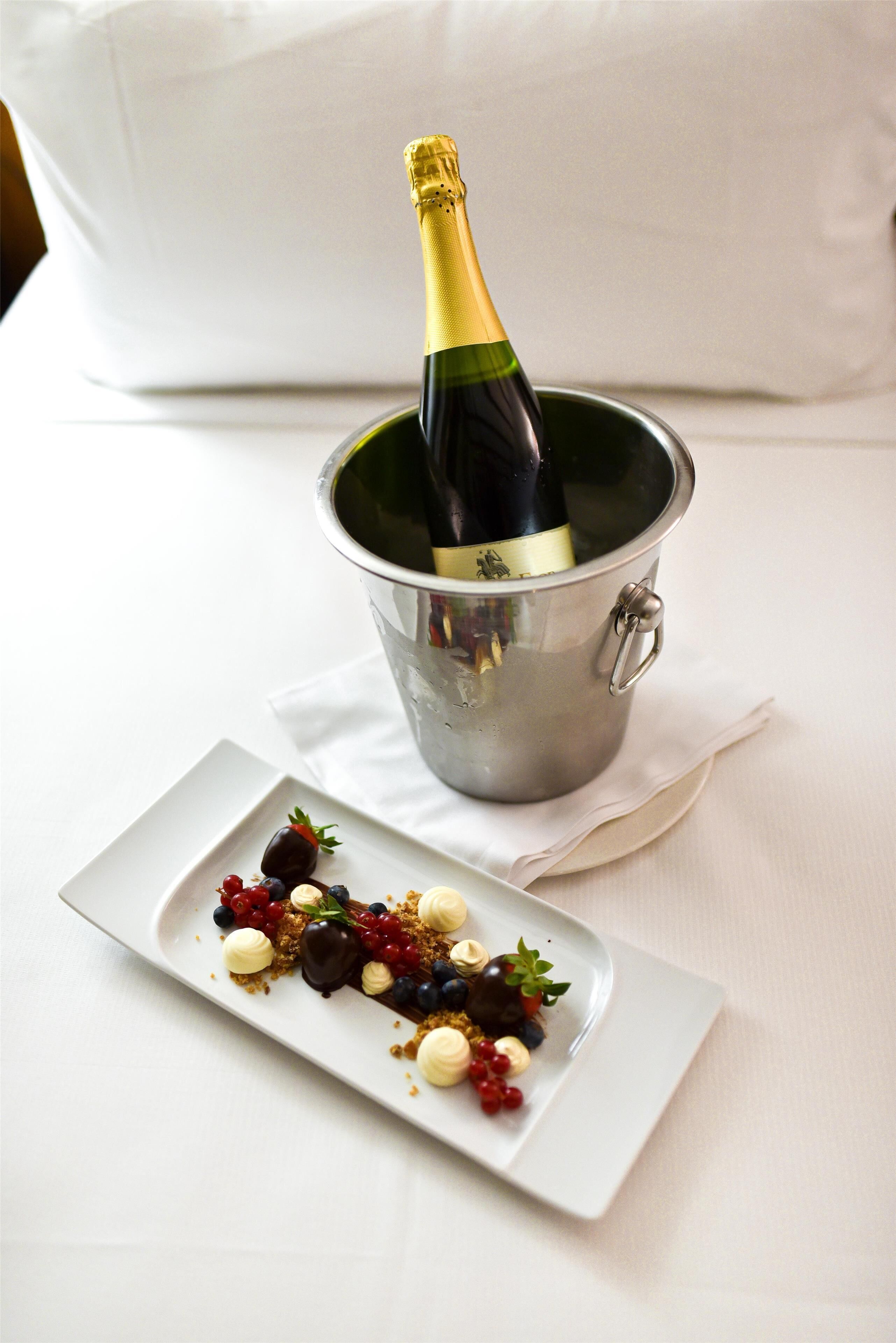 Una botella de champán en un balde de metal sobre un mantel blanco, acompañada por un plato rectangular de postre con bayas y crema.