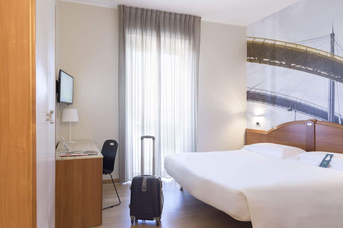 A hotel room with a bed, a desk with a lamp, a chair, a suitcase, and a large window with curtains.