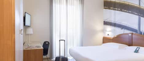 Habitación de hotel con cama, escritorio con lámpara, silla, maleta y un amplio ventanal con cortinas.