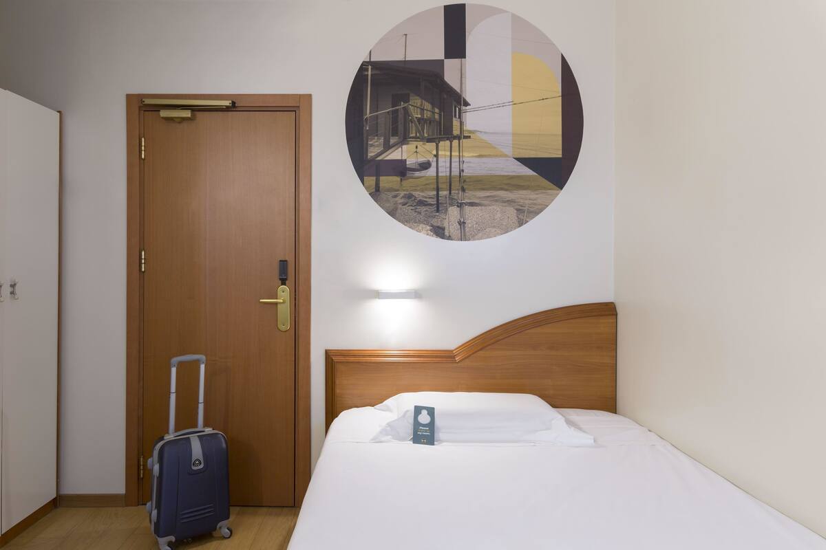 A hotel room with a wooden door, a bed with white bedding, and a circular wall art depicting a building and landscape.