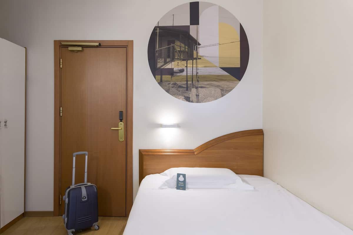 A hotel room with a wooden door, a bed with white bedding, a suitcase, and a circular wall art depicting a coastal scene.