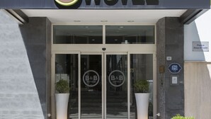 Der Eingang zu B&B Hotels mit Glastür, einem Hinweisschild auf den Standort und je einer Topfpflanze auf beiden Seiten.
