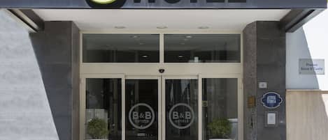 The entrance to B&B Hotels with a glass door, a sign indicating the location, and two potted plants on either side.