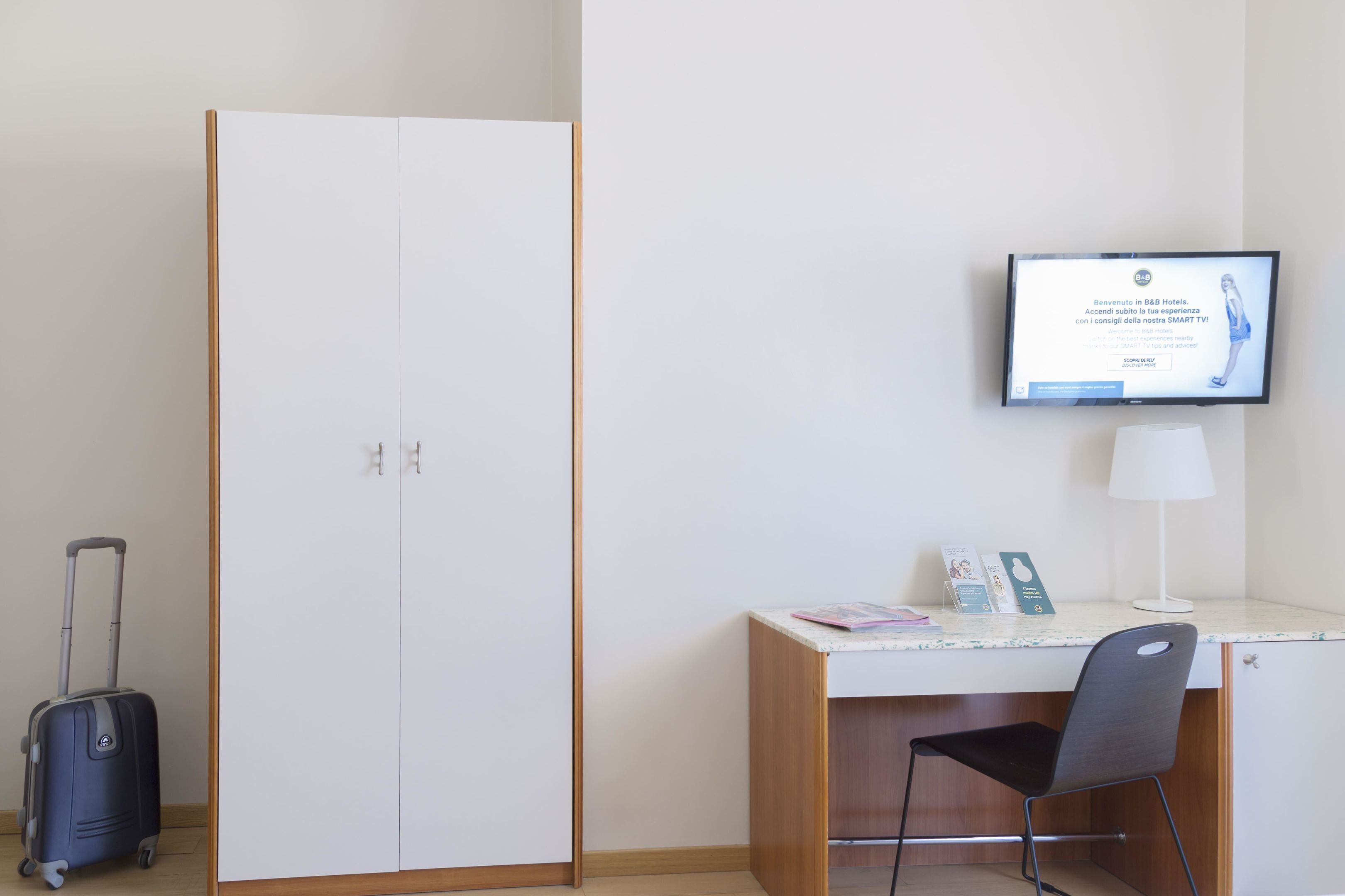 Een hotelkamer met een bureau, een stoel, een aan de muur bevestigde televisie en een koffer op de vloer.