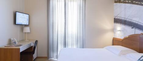 Habitación de hotel con cama, escritorio, silla, televisor y ventana con cortinas.