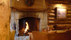 Interior de una cabaña de troncos con chimenea de piedra, pared de madera con un cuadro enmarcado y un rincón de estar acogedor.