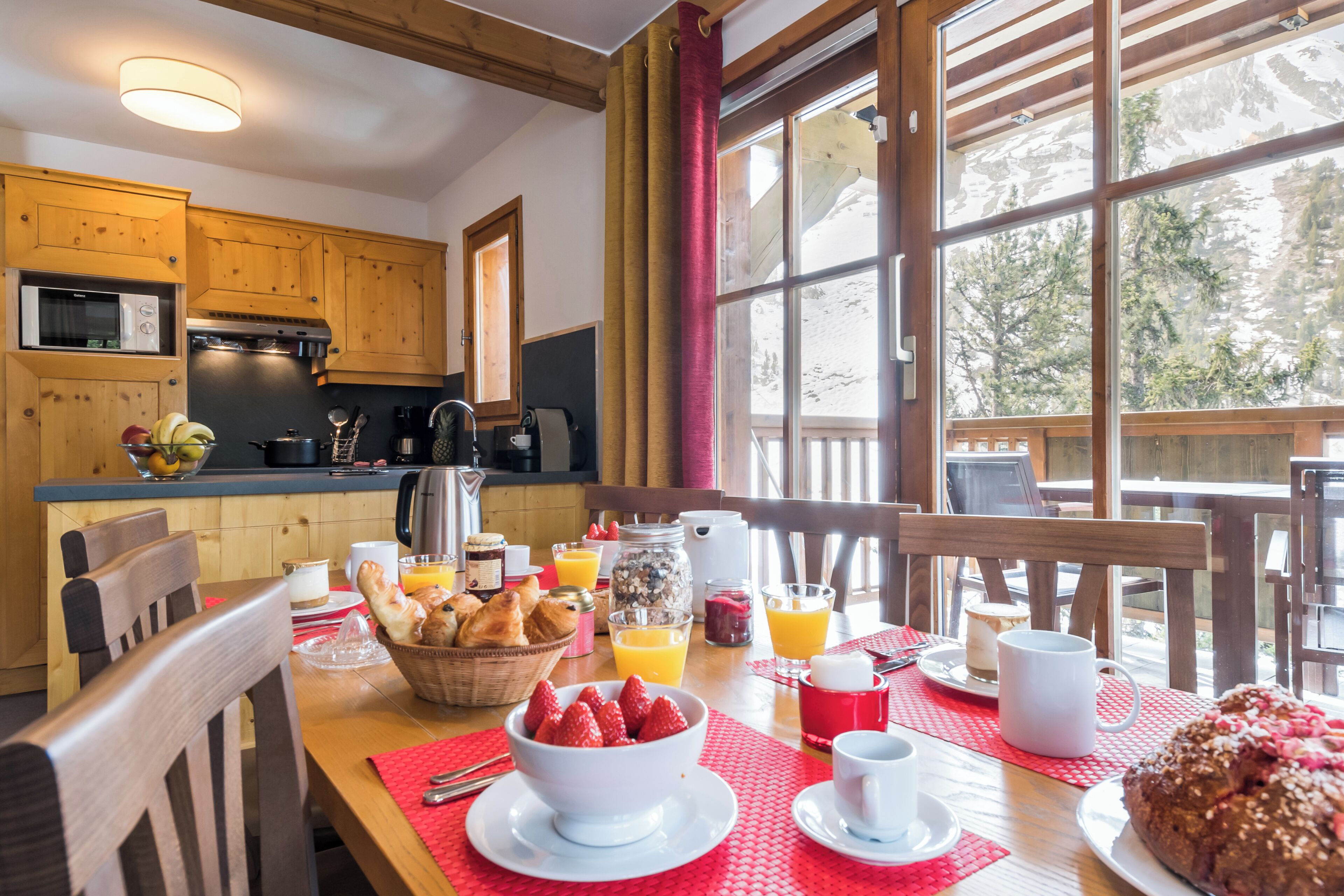 Ein gedeckter Frühstückstisch mit Brot, Obst und Getränken, Holzkunst, große Fenster mit Blick auf Bäume.