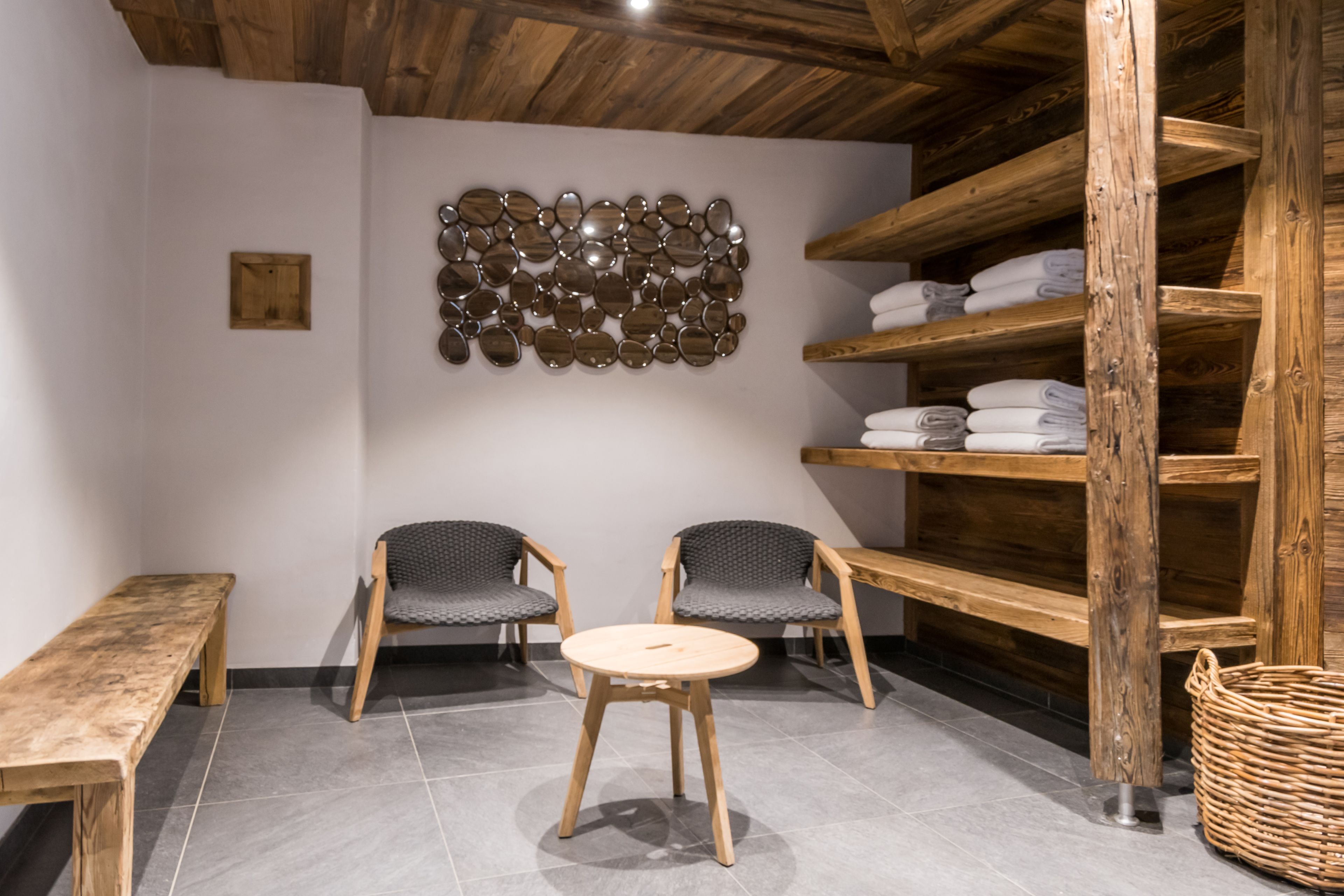 Habitació amb sostre de fusta, dues cadires tapissades de gris, una taula rodona de fusta, un mirall fixat a la paret, prestatges amb tovalloles plegades i una cistella de vimbles.