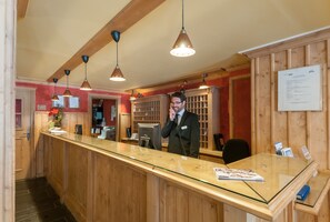 A reception area with a wooden counter, pendant lighting, and a man in a suit standing at the counter. - Pierre & Vacances Premium Arc 1950 Le Village (Bourg-Saint-Maurice)