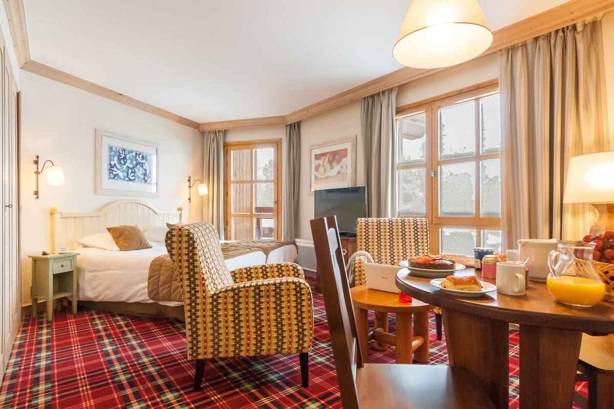 A hotel room with a bed, two patterned armchairs, a dining table with breakfast items, a TV, and large windows with curtains.