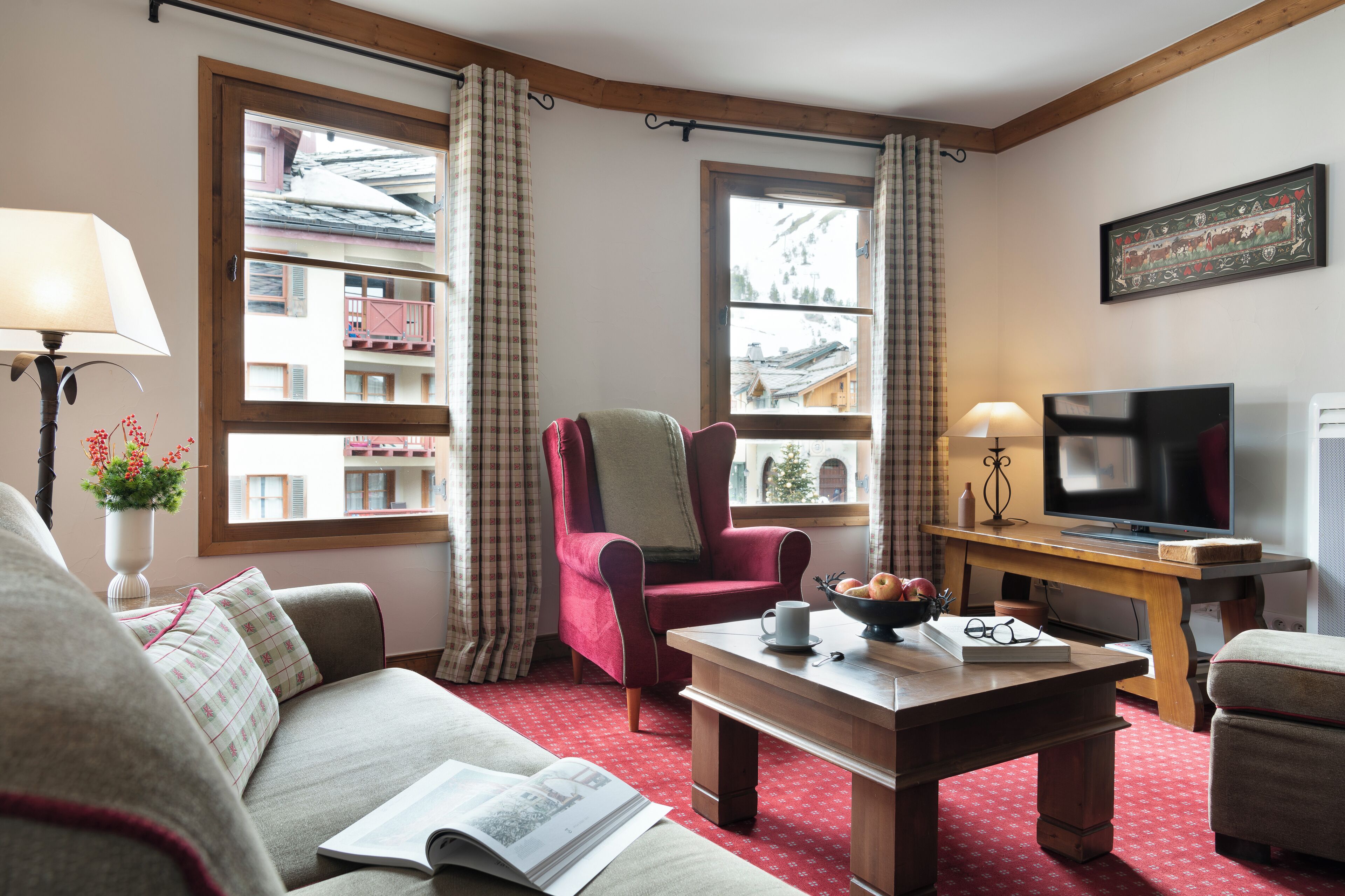 A cozy living room with a red armchair, a wooden coffee table, a television, and a large window with curtains.