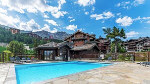 A traditional wooden chalet with a pool, surrounded by mountains and other buildings. - Pierre & Vacances Premium Arc 1950 Le Village (Bourg-Saint-Maurice)