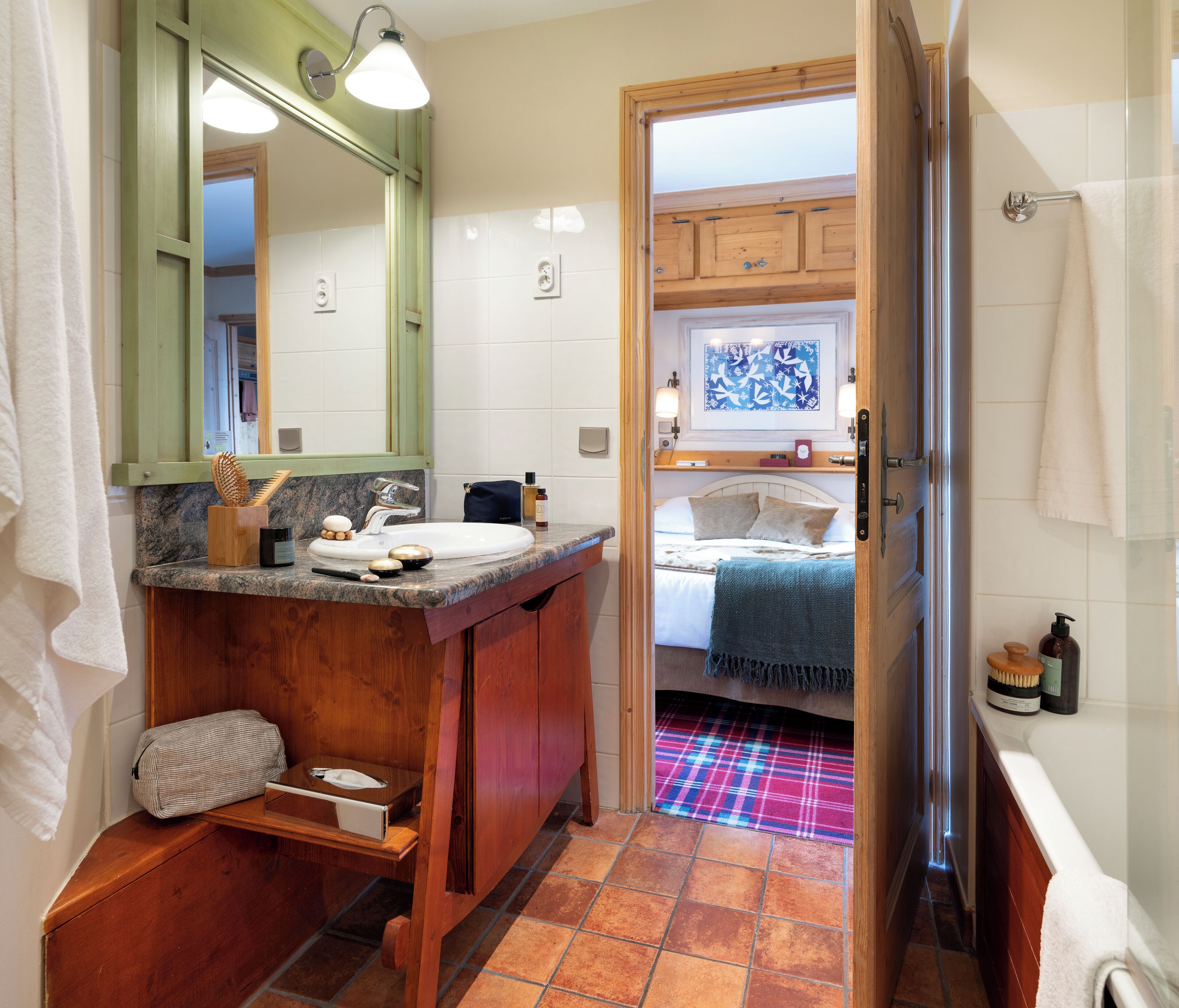 Un baño con tocador de madera, lavamanos y espejo. Se ve una puerta que da a un dormitorio con cama y ventana.