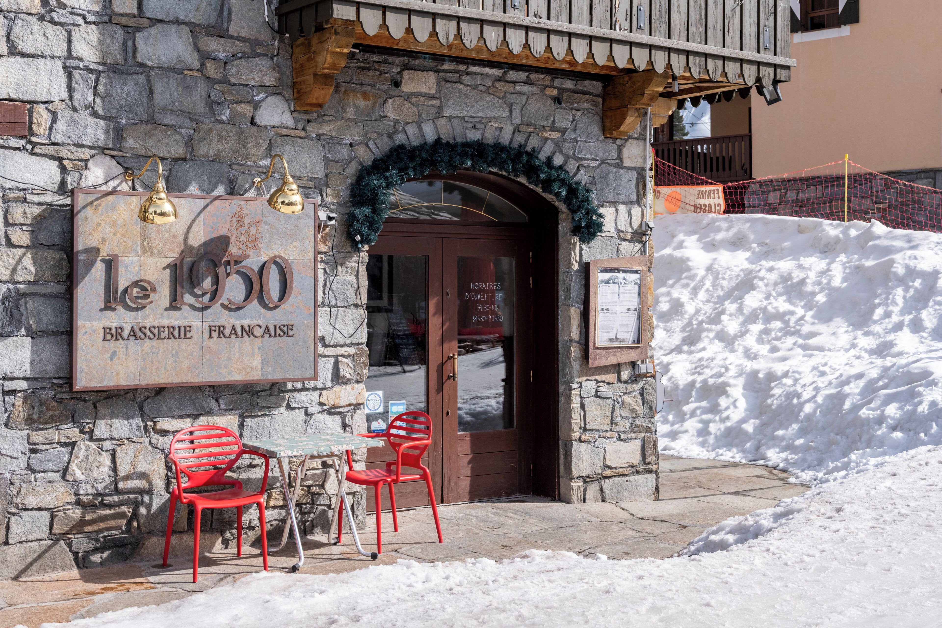 A stone building with a sign reading 'le 1950 BRASSERIE FRANCAISE,' outdoor seating with red chairs, and a snow-covered ground.