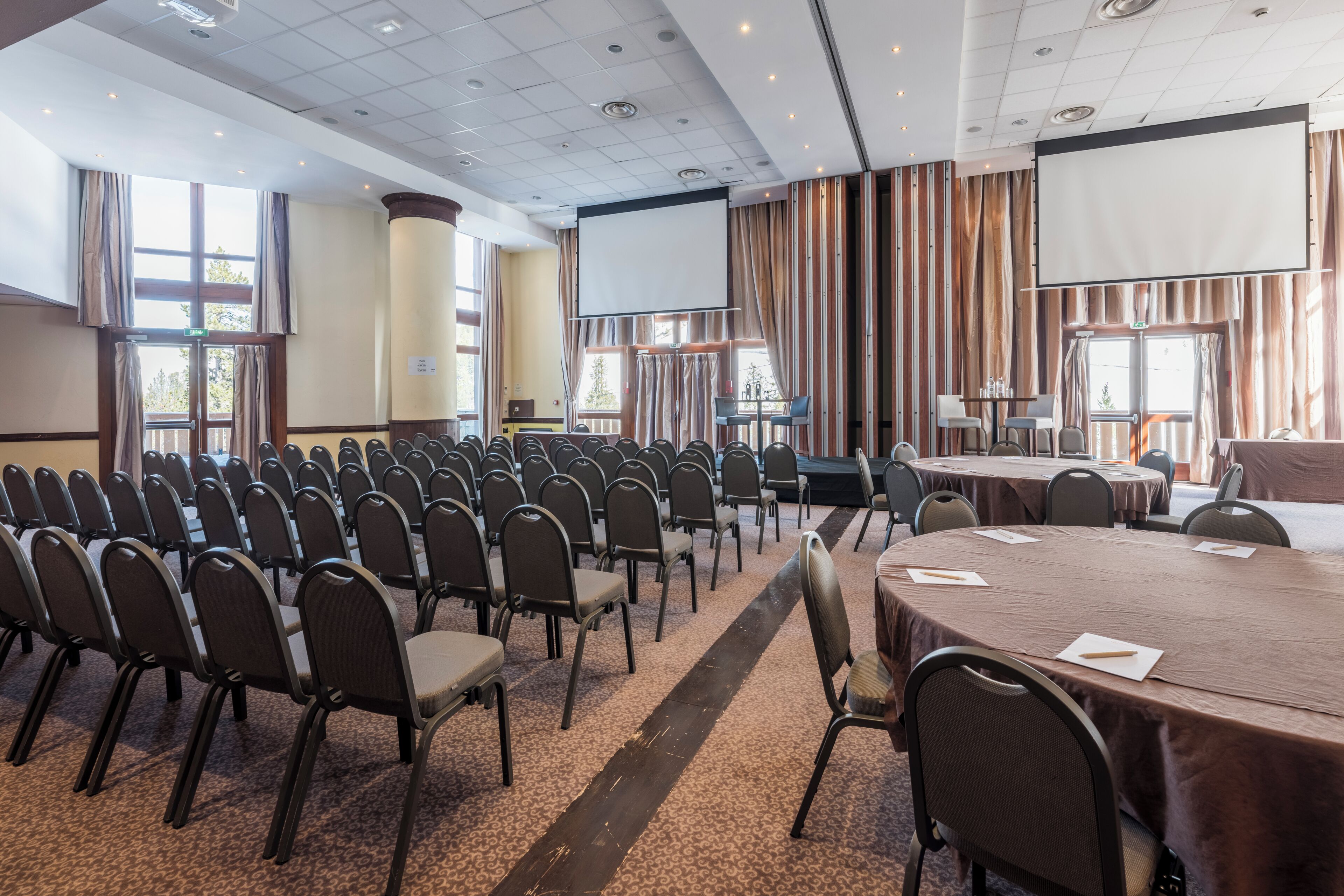 A conference room with rows of chairs, a large table, a projector screen, and a view of the outdoors through large windows.