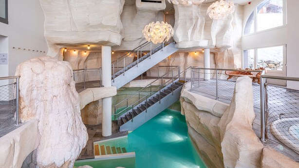 Una piscina interior de varios niveles con una escalera central, rodeada de estructuras que imitan rocas y un aplique colgante.