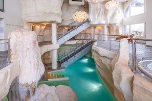 A multi-level indoor pool with a central staircase, surrounded by rock-like structures and a hanging light fixture. - Pierre & Vacances Premium Arc 1950 Le Village (Bourg-Saint-Maurice)