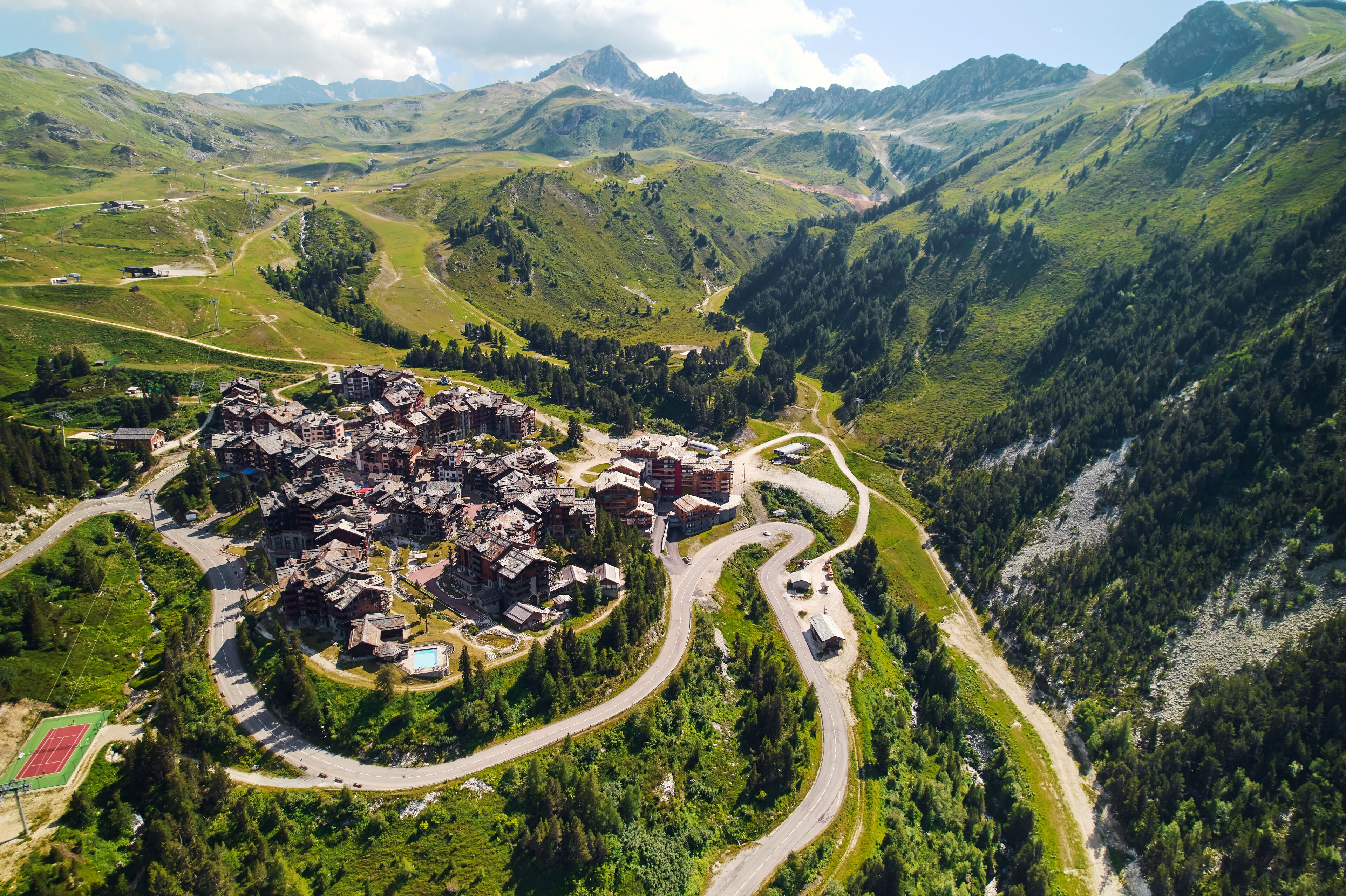 Une vue aérienne d’un village avec des routes sinueuses, des collines verdoyantes et un ensemble de bâtiments dont l’architecture est un mélange de styles traditionnels et modernes.