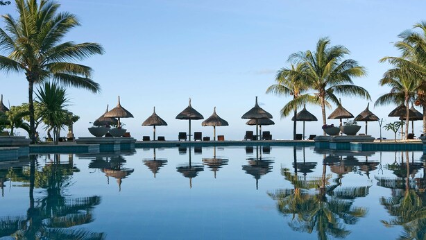 A poolside area with lounge chairs and thatched umbrellas, surrounded by palm trees.